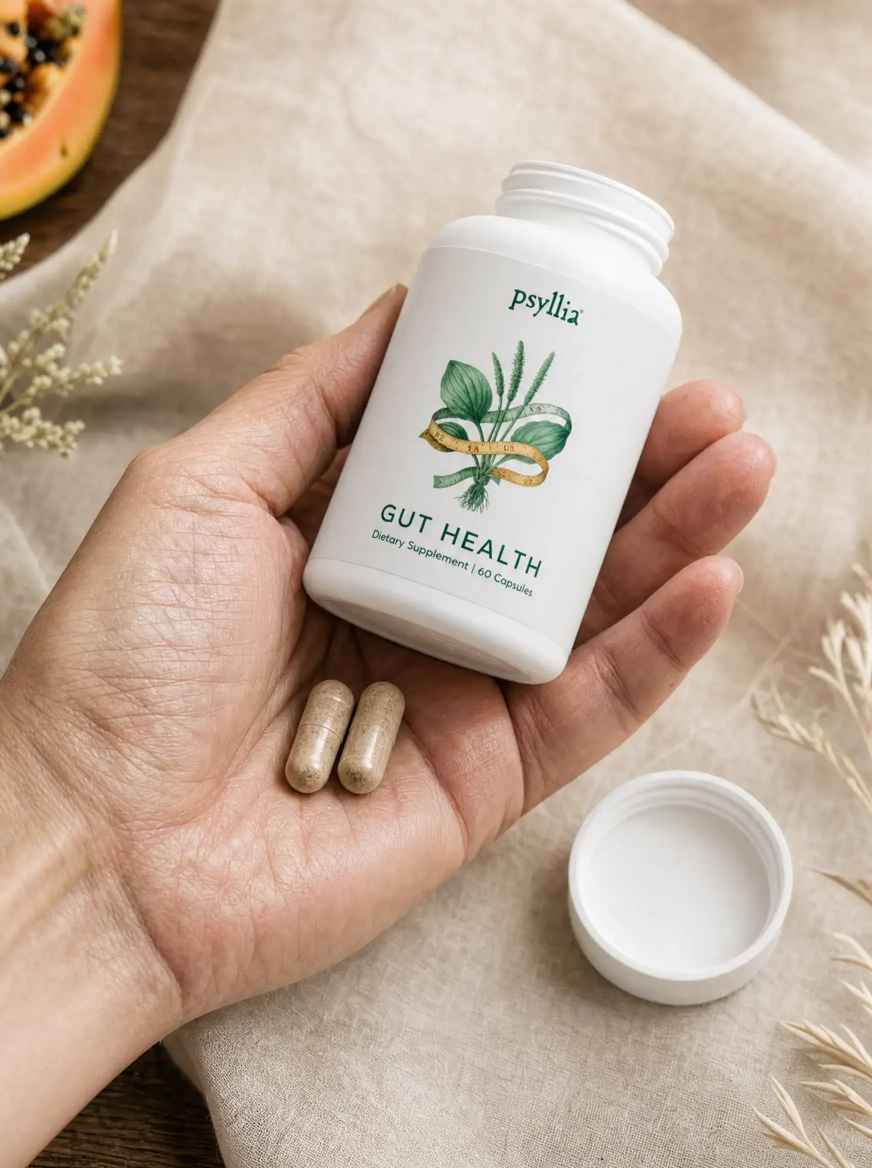 A hand holding the open Veradine bottle with two capsules resting in the palm. The white cap is set down on warm linen, with papaya and dried grass in soft focus.